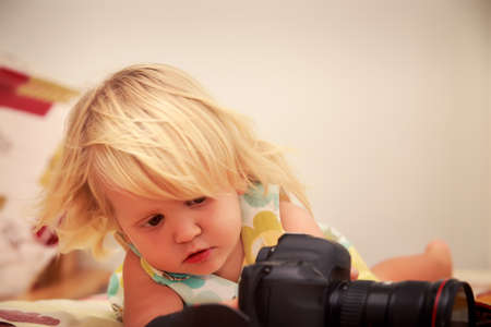 little blonde girl in colorful dress looks with interest into object-glass of photo camera on sofa against white wallの写真素材