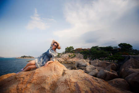 young brunette girl in short grey frock with lace and sunglasses lies on rock against stones plants and seaの写真素材