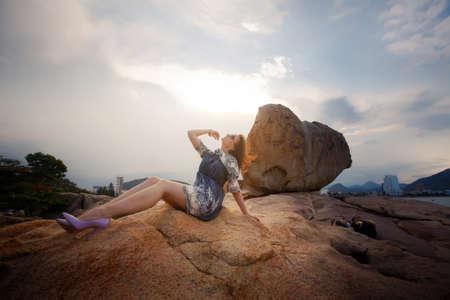 young brunette girl in short grey frock with lace and sunglasses sits on rock and looks into sky against sky and cloudsの写真素材