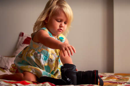 little blonde girl in colorful dress looks with interest into object-glass of photo camera on sofa against white wallの写真素材