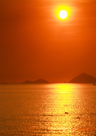 people swim in sea against sun white disk among red sky above sea and islands at sunrise