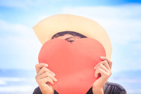 closeup European guy in nice white hat hides face behind hand-made large red heart against seaの写真素材
