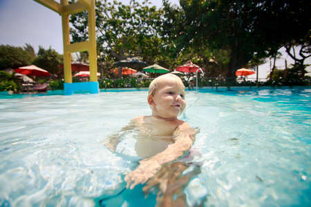 small blonde girl swims in shallow transparent water of hotel swimming pool against colourful parasolsの写真素材