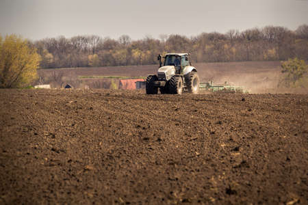tractor on big wheels cultivates and raises great dust on ploughed soil against village and forestの写真素材