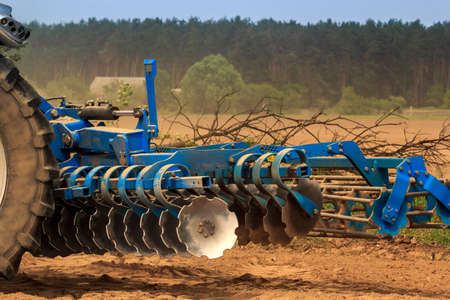 cultivator stands on ground road by ploughed field against village and forestの写真素材