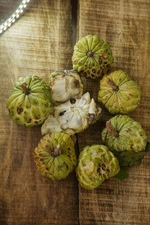 top view picture of broken ripe sugar-apple with brown seed among unbroken fruits on wooden table...