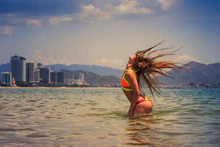 blonde slim female gymnast in bikini stands half body in sea and shaken head lifts hair up over head against cityの写真素材
