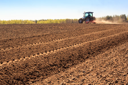 distant cultivator on big wheels operates on large ploughed field raises dust behind