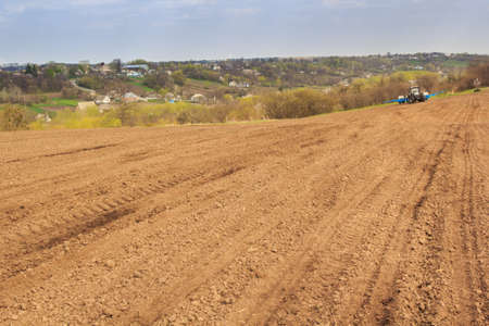 tractor in brown ploughed field in idyllic spring against distant forest and Ukrainian village on hillsの写真素材