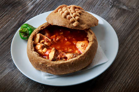 tasty red soup with pieces of fish in original bowl made from homemade brown bread on white plate with green pepper against dark wooden tableの写真素材