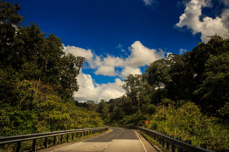 asphalt road along wooded hill out of front bus window with driver side mirror against cloudy blue...