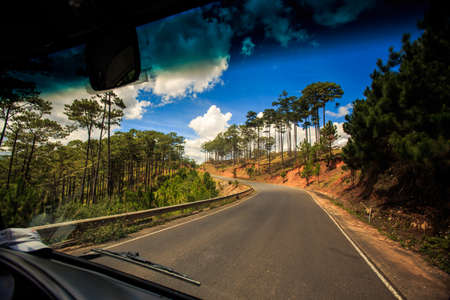 sunny highway along brown hill and tropical plants and driver side mirror against cloudy blue sky out of bus windowの写真素材