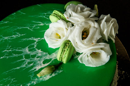 closeup homemade mousse cake decorated with green mirror glaze, white roses, coconut chips and...