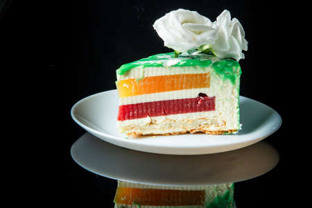 side view of piece of tasty green-glazed cake with yellow and red jelly inter-layer and decorated with flower on top served on white plateの写真素材
