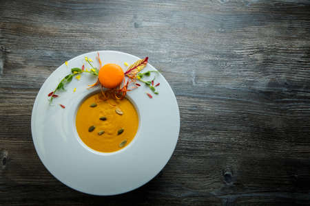 top view of fresh pumpkin cream soup with seeds and orange macaroon decoration on deep white plate on wooden backgroundの写真素材