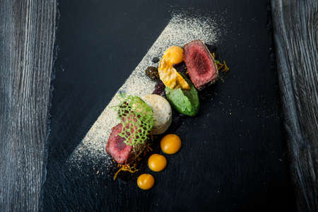 top view on two pieces of roasted meat decorated with puree and sauce on black plate on wooden table in restaurantの写真素材