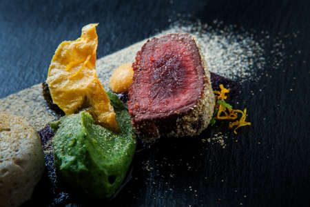macro two pieces of roasted meat decorated with puree and sauce on black plate on wooden table in restaurantの写真素材