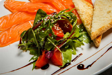 Rucola and tomato salad served with salmon slices and toasted bread on white restaurant plateの写真素材