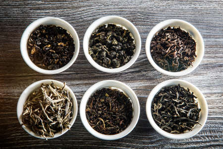 top view of different kinds of tea leaves in six white small round vessels on wooden table backgroundの写真素材