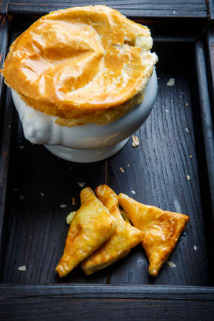 closeup big white soup pot with bread on the top and three puff pastry cakes triangles served n black wooden trayの写真素材
