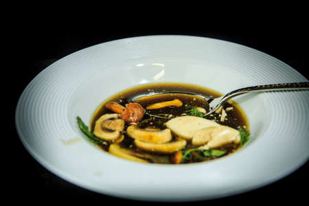 closeup vegetarian soup with mushrooms, mozzarella cheese, and dill served in restaurant white plate and spoon on black backgroundの写真素材