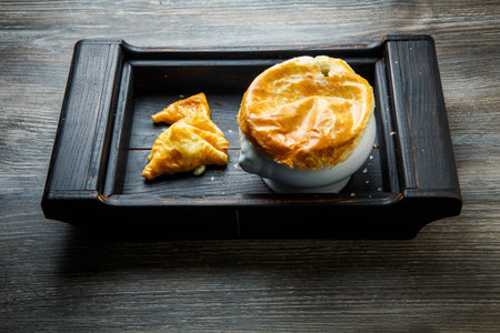 big white soup pot with bread on the top and three puff pastry cakes triangles served on black wooden trayの写真素材
