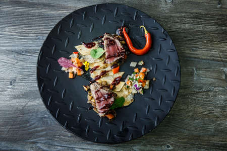 top view two pieces of medium raw grilled meat decorated with red pepper and sauce served with potato chips and vegetable salad on black restaurant plate.の写真素材