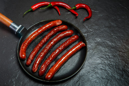 top view on round pan with five long fried hunting sausages served with three red chili pepper on...