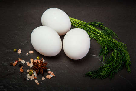 Closeup three white chicken eggs served with bunch of fresh green dill and different spices on black backgroundの写真素材