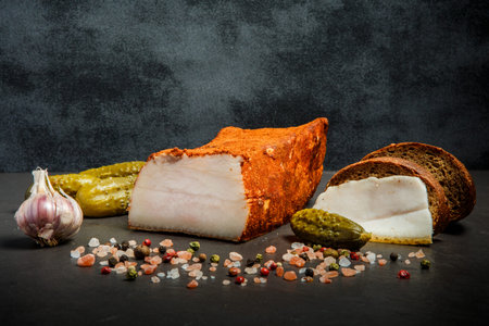 Sliced raw lard in red pepper served with garlic, aromatic spices, rye bread and pickled cucumbers on black backgroundの写真素材