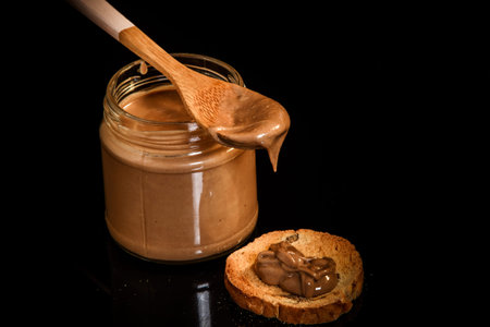 top view closeup creammy peanut butter in glass jar, wooden spoon, and small cracker on black...