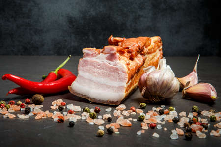 big chunk of salted bacon served with whole garlic, pink salt, aromatic spices, red chili pepper on black backgroundの写真素材