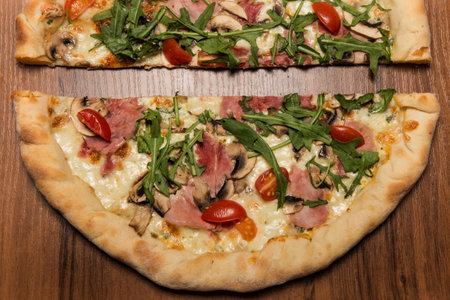 top view closeup two halves of pizza with cheese, arugula, sliced ham, mushrooms and tomatoes on...