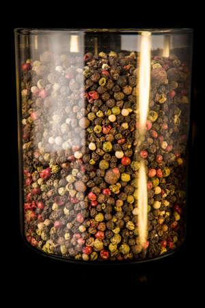 closeup tall glass jar with assorted peppercorns black, red, green, white and allspice peppercorns on black backgroundの写真素材