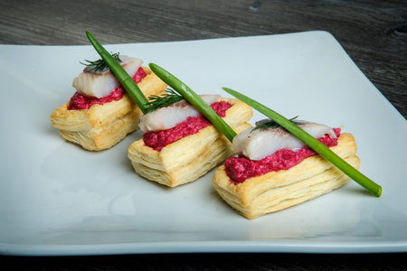 closeup snacks with red sauce, herring pieces, and green onion served on restaurant white plate on...