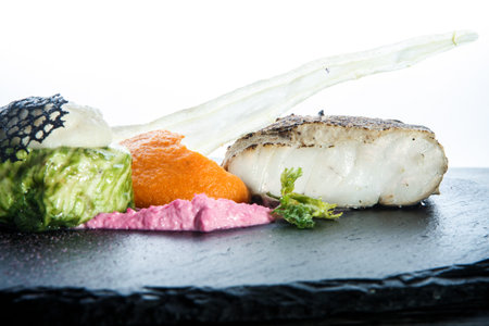 closeup roasted white fish with carrot puree and vegetables served on black plate against white...