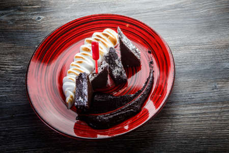 delicious sliced homemade brownie cake with baked white cream and chocolate sauce on red restaurant plate on wooden tableの写真素材