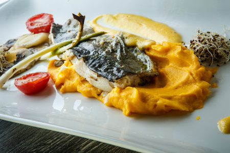 closeup yellow potato puree decorated with baked fish fillet and vegetables on modern white plate on...