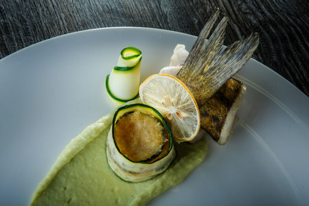 top view closeup finely decorated fish roll with sliced zucchini and green puree garnish on white...