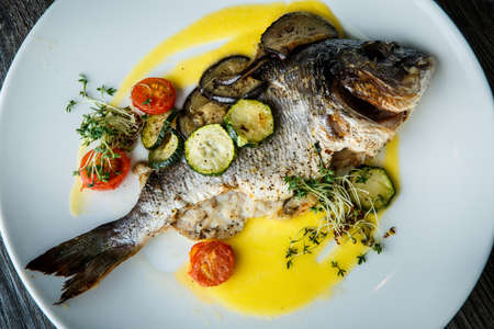 closeup delicious baked fish served with sliced baked vegetables on white round plate on dark tableの写真素材