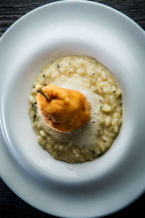 vertical view closeup boiled barley porridge decorated with breaded whole pear served in deep white restaurant plate on wooden tableの写真素材