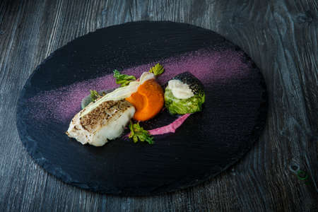 deliciousroasted white sea fish with carrot puree and vegetables served on black plate on dark wooden tableの写真素材