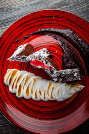 vertical view on tasty sliced brownie cake with baked white cream and chocolate sauce on red restaurant plate on wooden tableの写真素材