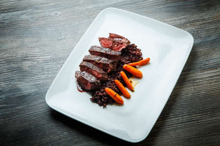 top view on delicious dish of barbecued sliced meat served with wild rice and grilled carrot garnish on modern white plate on wooden tableの写真素材