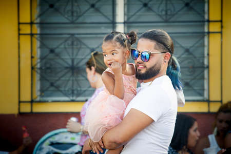 Concepcion De La Vega, DOMINICAN REPUBLIC - FEBRUARY 09, 2020: dominican man in blue glasses carries small girl in princess dress on his hands on February 9 in Concepcion De La Vegaのeditorial素材