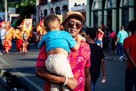 Concepcion De La Vega, DOMINICAN REPUBLIC - FEBRUARY 09, 2020: adult man in dark glasses carries little son walking by city street at dominican carnival on February 9 in Concepcion De La Vegaのeditorial素材
