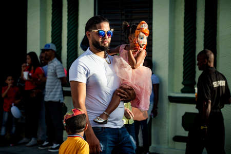 Concepcion De La Vega, DOMINICAN REPUBLIC - FEBRUARY 09, 2020: adult man in blue glasses carries little girl in vivid dress and mask on his hands on February 9 in Concepcion De La Vegaのeditorial素材