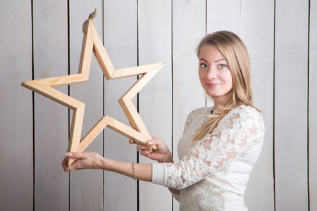 smiling slim blonde girl looks at camera holding in hands small beige wooden star shaped decoration...