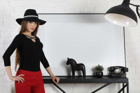 Attractive young fashionable girl posing in red dress and black hat indoors. Space for your text or logoの写真素材