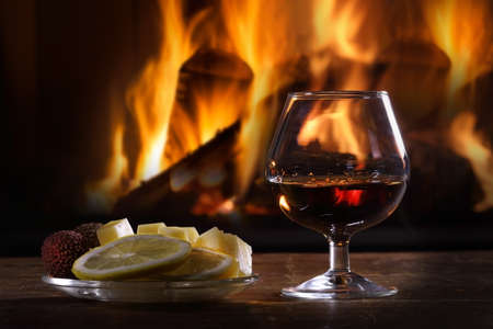 Still life of a glass of brandy and fruit with cheese on the background of a lit fireplaceの写真素材